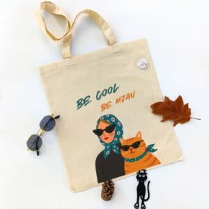 Ceger Be Mjau sa ilustracijom žene i narandžaste mačke sa naočarima za sunce i maramama, uz natpis „Be cool, be mjau“. Poklon za ljubitelje mačaka i stila.
